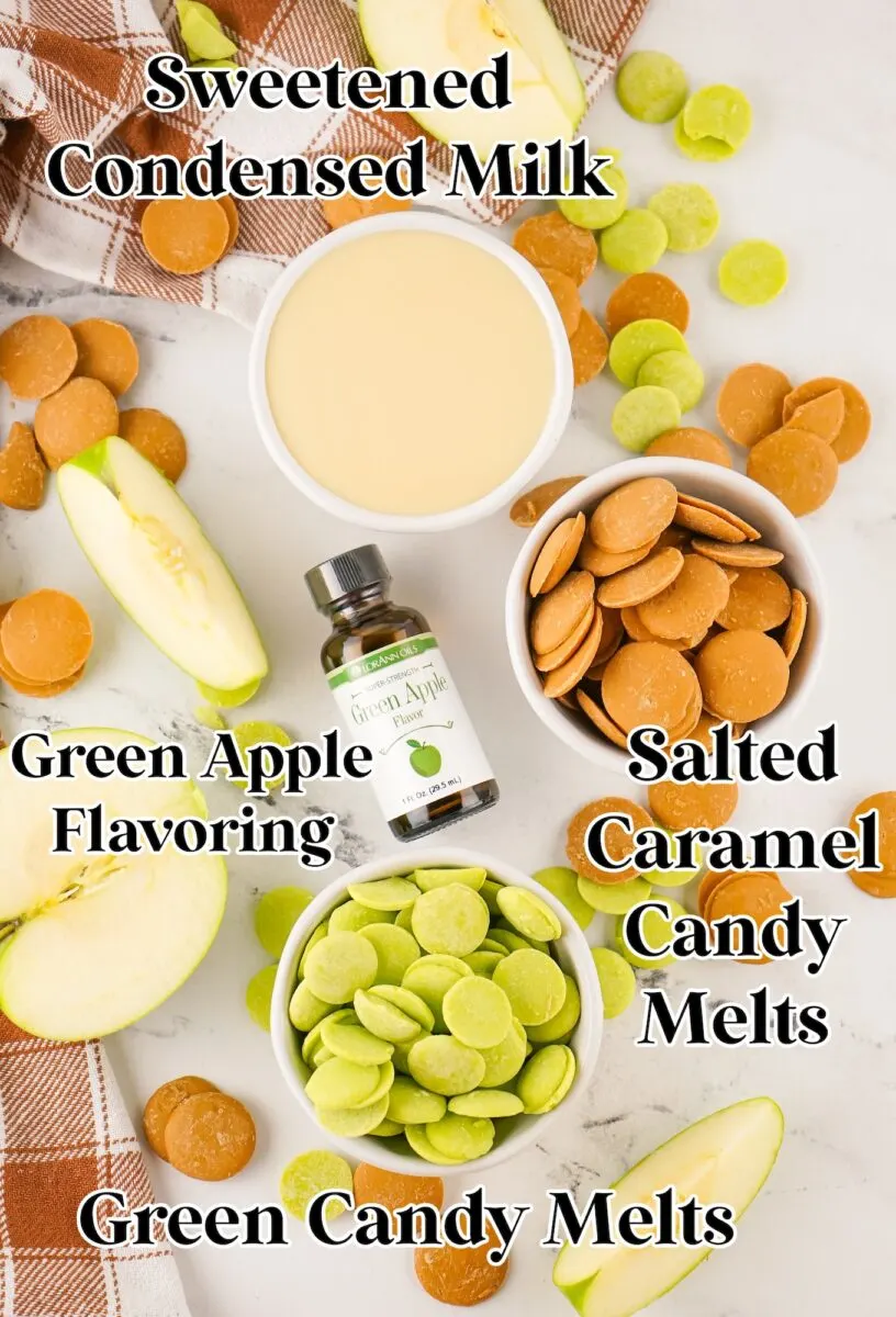 Flat lay of ingredients for caramel apple fudge, including sweetened condensed milk, green apple flavoring, green and salted caramel candy melts, apple slices, and a plaid cloth on a marble surface.