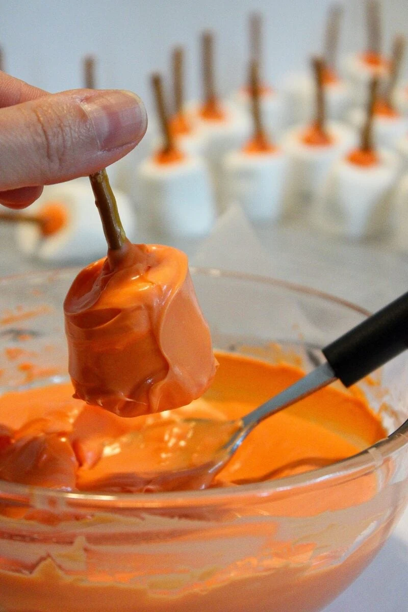 A hand holds a pumpkin patch marshmallow on a pretzel stick, dipping it into a bowl of orange coating, with finished treats in the background.