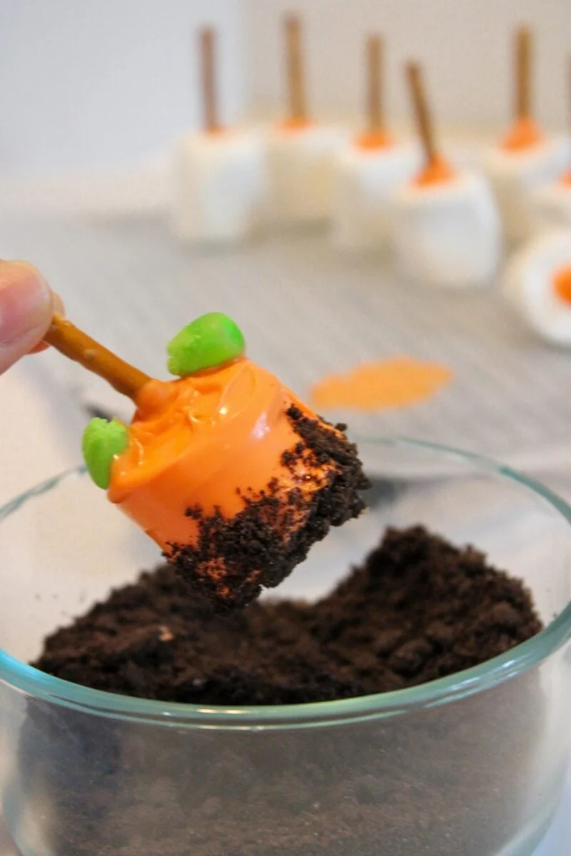 A hand dips a pumpkin patch marshmallow, coated in orange candy and topped with a pretzel stick and green candy, into a bowl of crushed chocolate cookies. More decorated marshmallows are in the background.