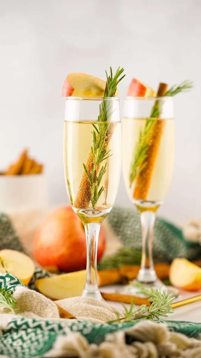 Two champagne flutes filled with a light drink, garnished with apple slices, rosemary sprigs, and cinnamon sticks, set on a table with apples and a green cloth.
