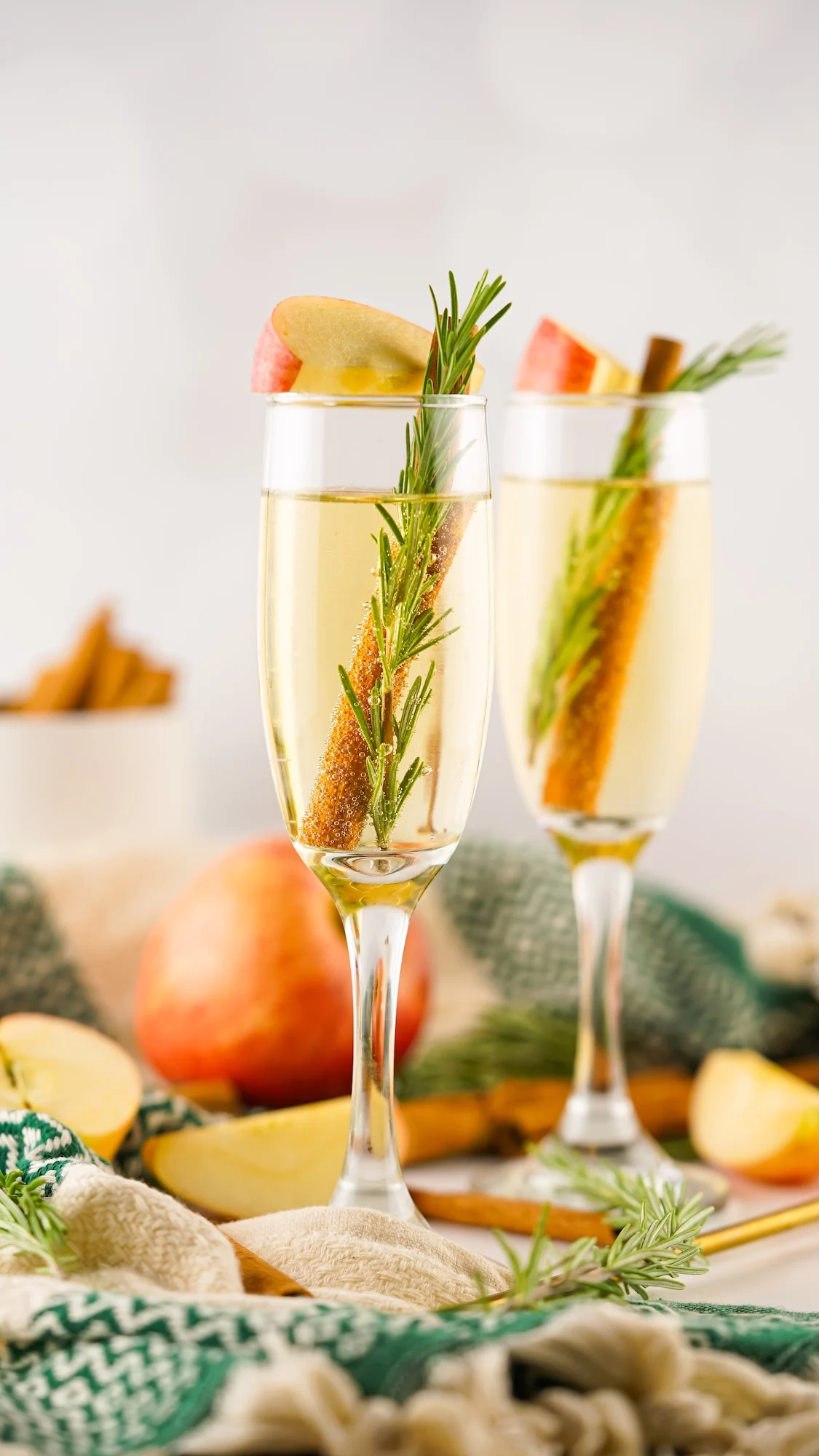 Two champagne flutes filled with a light drink, garnished with apple slices, rosemary sprigs, and cinnamon sticks, set on a table with apples and a green cloth.