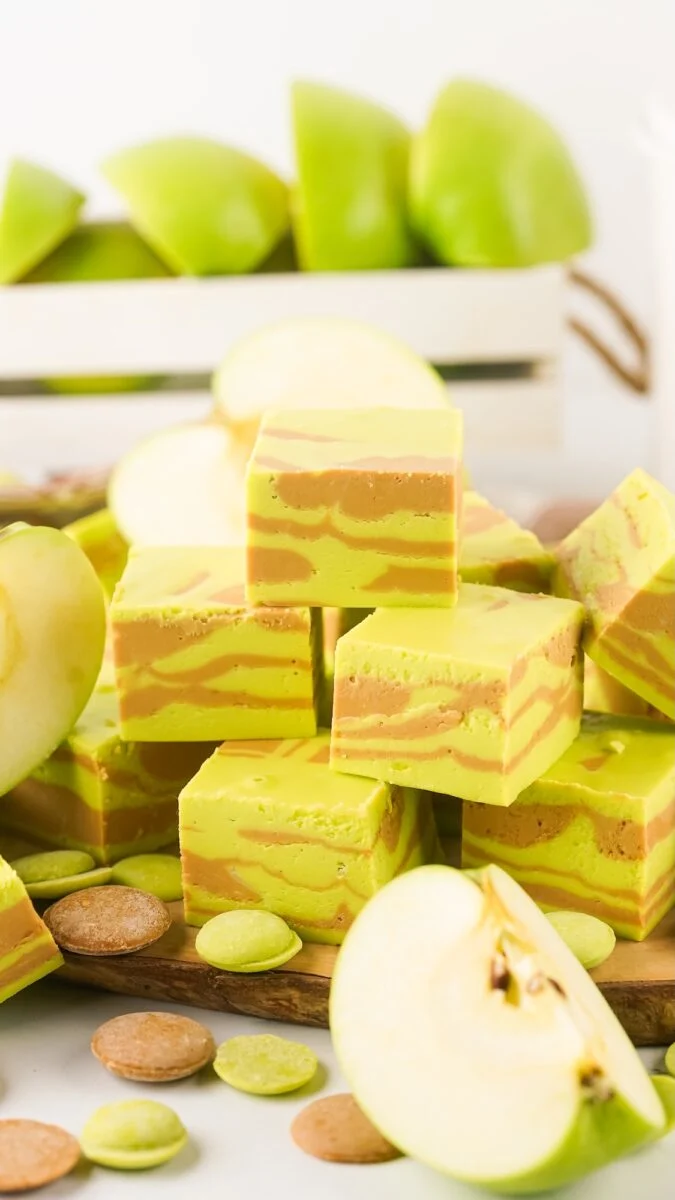 Squares of green and tan caramel apple fudge are stacked on a board, surrounded by sliced green apples and baking wafers, with a crate of whole green apples in the background.