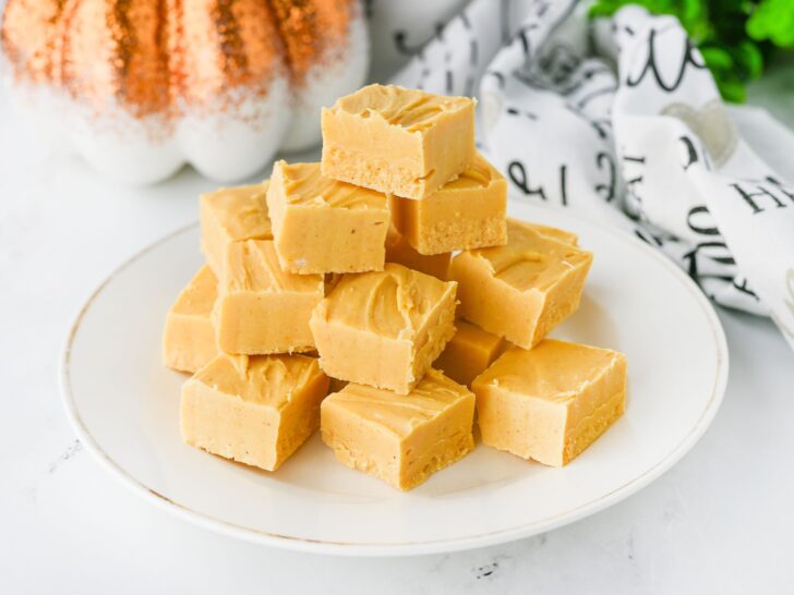 A white plate with a stack of square pieces of light brown fudge. A decorative pumpkin and patterned cloth are in the background.