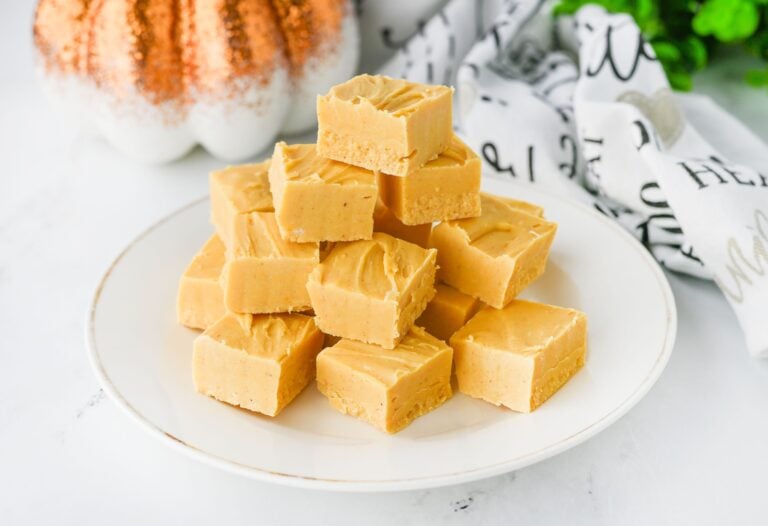 A white plate with a stack of square pieces of light brown fudge. A decorative pumpkin and patterned cloth are in the background.