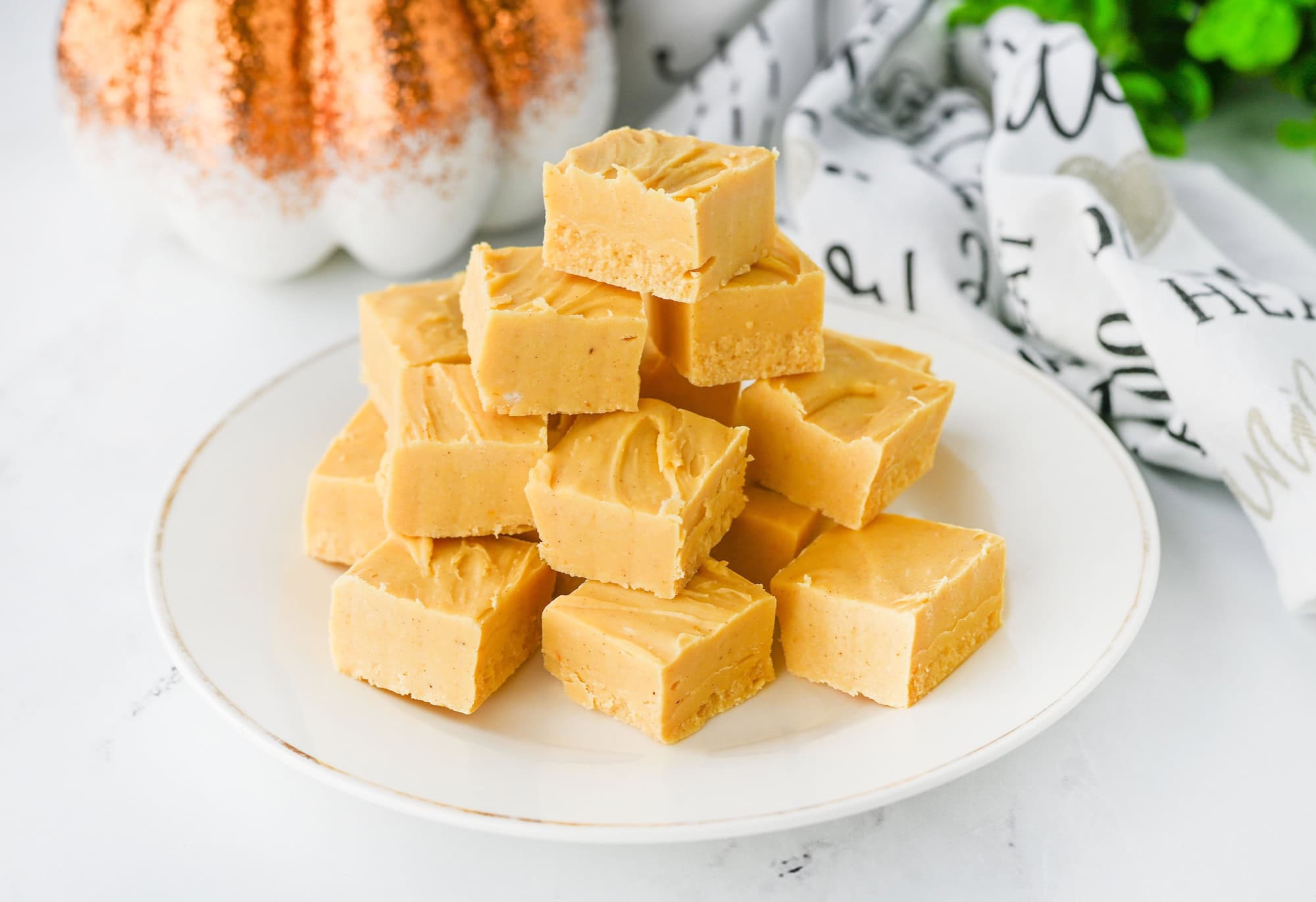 A white plate with a stack of square pieces of light brown fudge. A decorative pumpkin and patterned cloth are in the background.