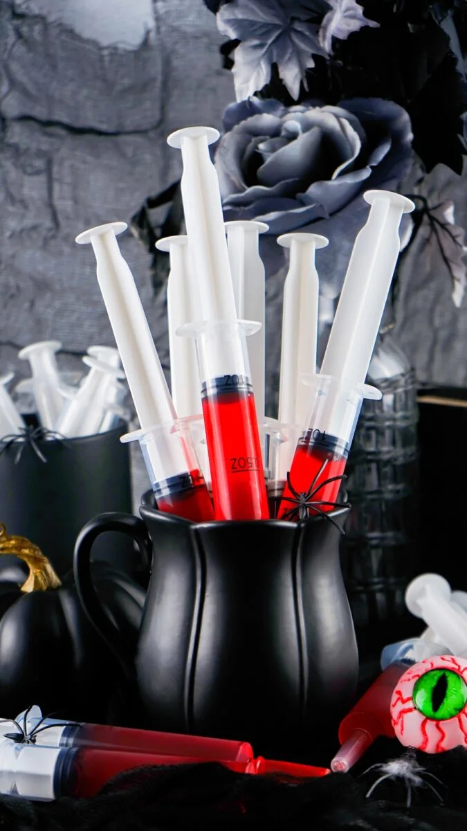 A black pitcher holds several syringe jello shots filled with red liquid, surrounded by Halloween-themed decorations including faux flowers and a fake eyeball.