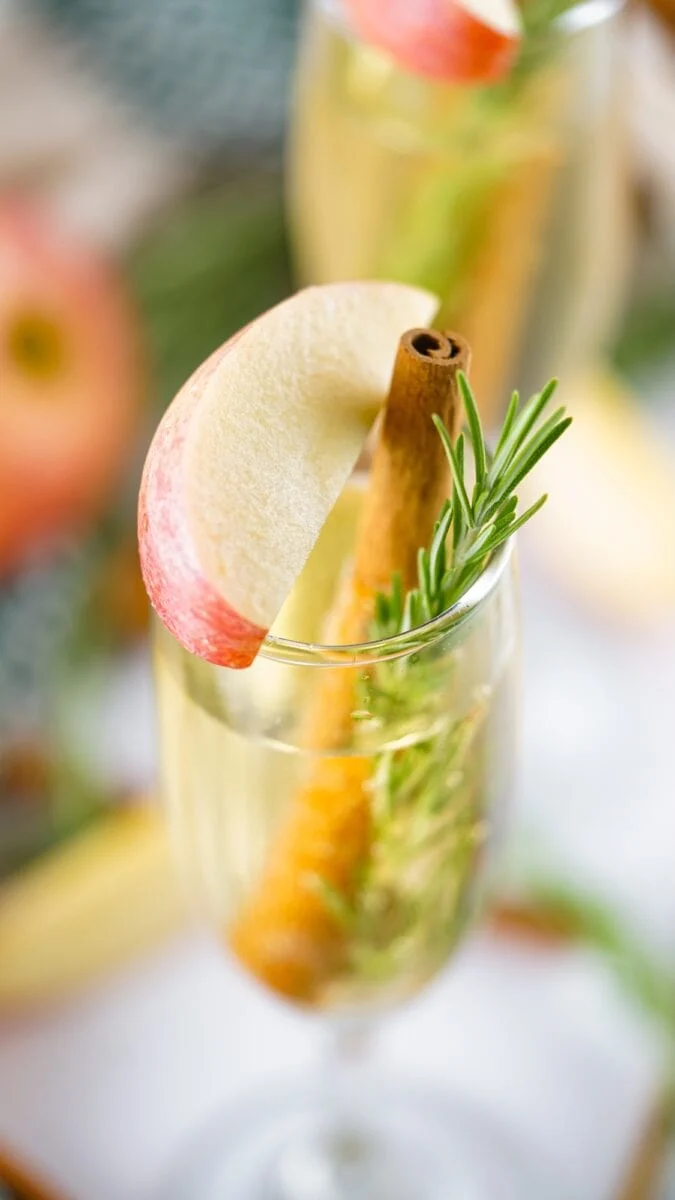 A glass of sparkling drink garnished with a cinnamon stick, a sprig of rosemary, and an apple slice.