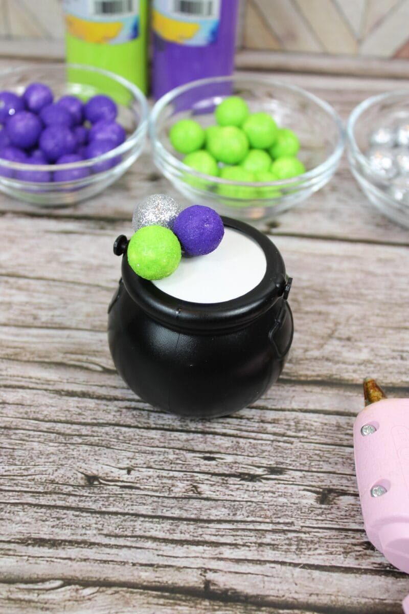 A small black cauldron filled with white material, topped with green, purple, and silver balls; bowls of colorful balls and a hot glue gun are nearby on a wooden surface.