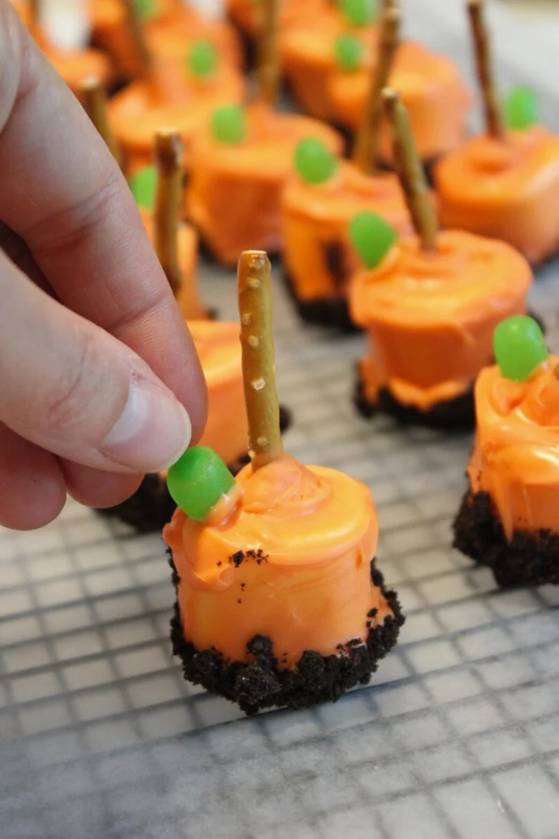 A hand places a green candy onto an orange, frosting-covered treat with a pretzel stick and cookie crumb base, resembling a pumpkin patch marshmallow on a cooling rack.