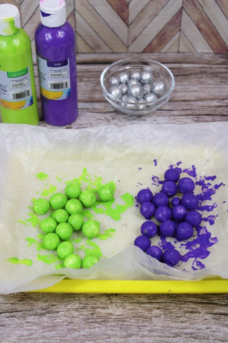 Two groups of painted balls, one green and one purple, are drying on wax paper in a tray. Bottles of paint and a bowl of silver balls are in the background.