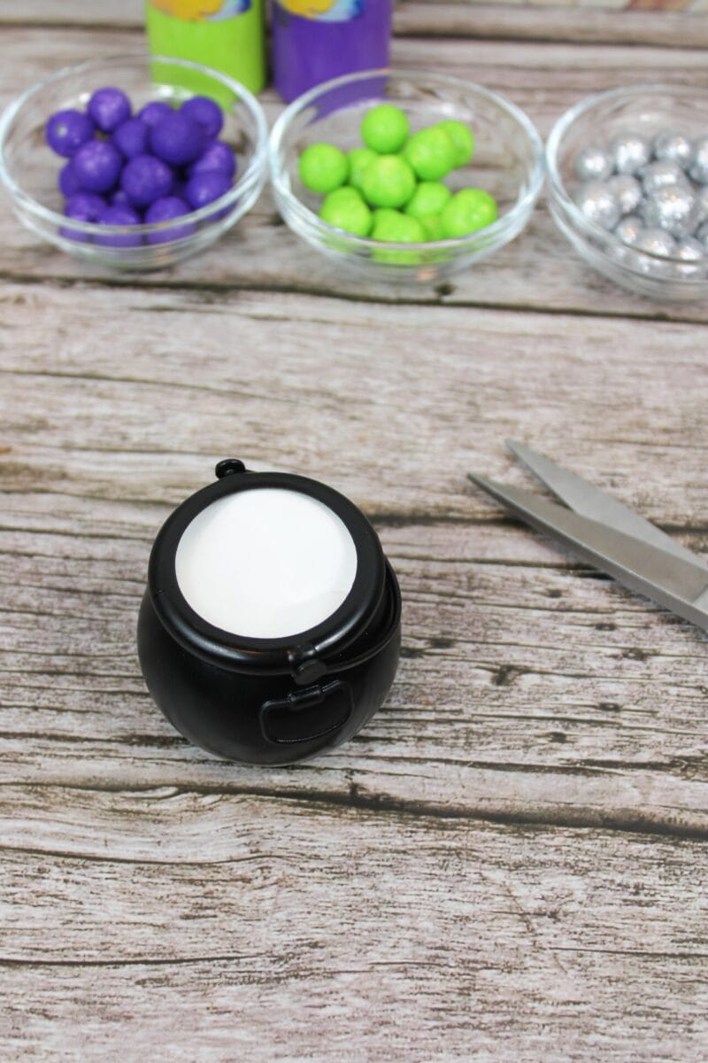 A small black cauldron filled with white liquid sits on a wooden surface near scissors and three bowls of colorful candy balls—purple, green, and silver.