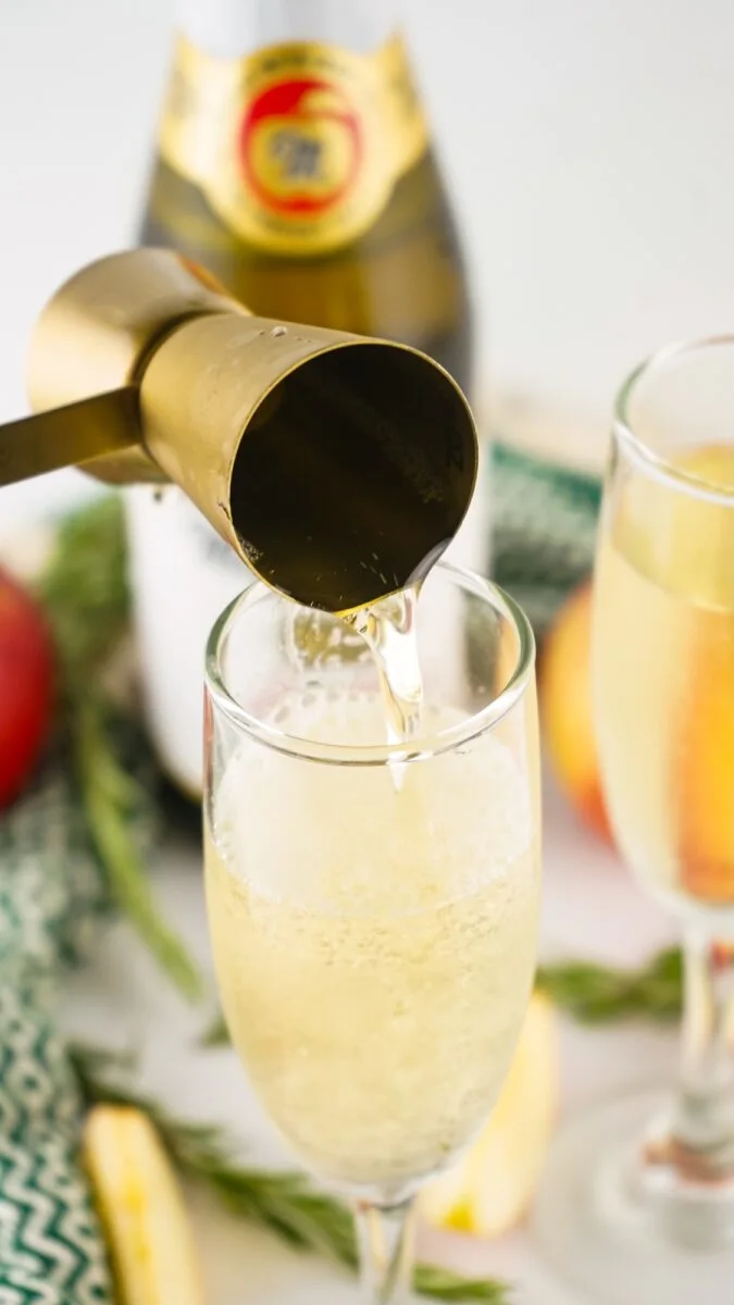 A brass jigger pours sparkling wine into a champagne flute, with another filled flute, a bottle, and fruit in the background.