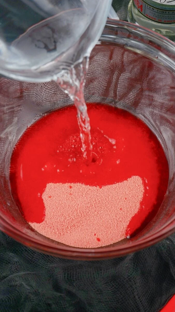 Water being poured into a glass bowl containing red powder and granules, likely for mixing a drink or preparing syringe jello shots for a fun party treat.