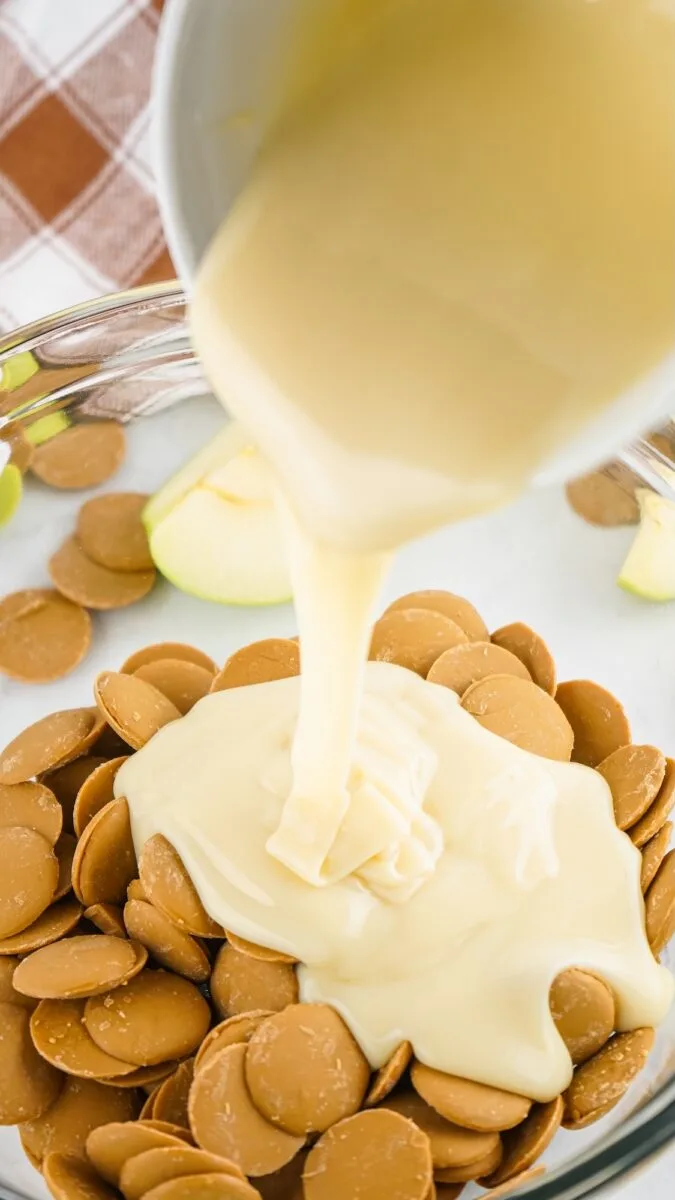 White melted chocolate is poured over caramel candy melts in a glass mixing bowl, creating the perfect base for caramel apple fudge, with sliced green apples visible nearby.