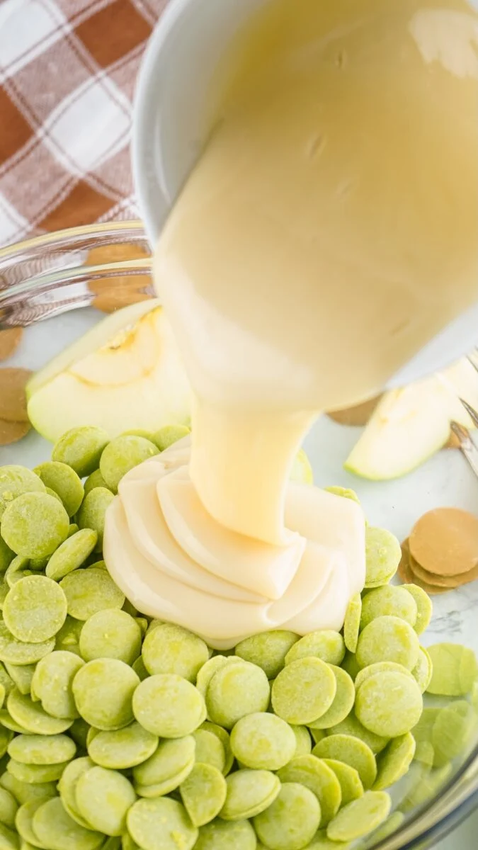Sweetened condensed milk is poured onto green candy melts in a glass bowl, with apple slices and caramel pieces nearby—perfect for making delicious caramel apple fudge.