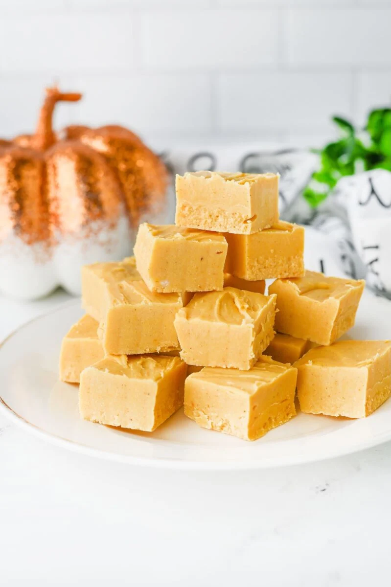 A plate stacked with square pieces of light brown fudge sits on a white surface, with a decorative pumpkin and green plant in the background.