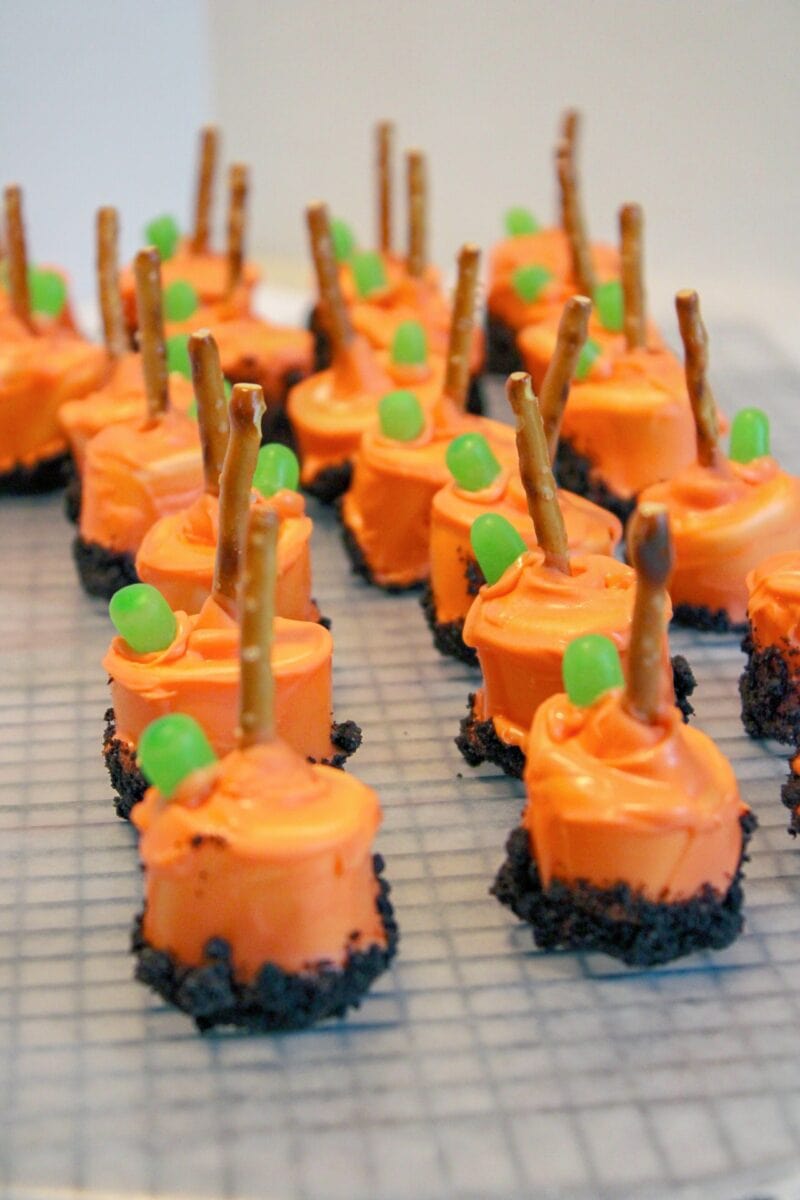 Rows of pumpkin patch marshmallow treats with orange frosting, crushed cookie bases, pretzel stick handles, and green candy decorations are arranged on a cooling rack.
