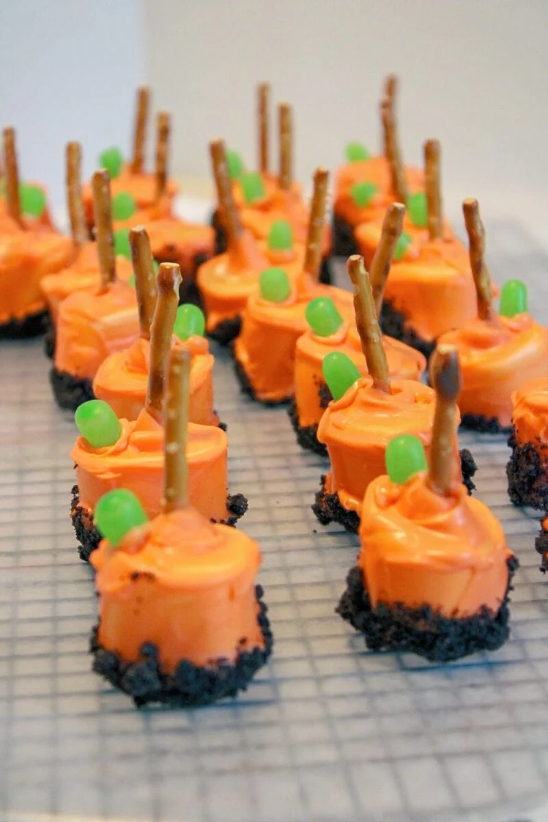 Rows of pumpkin patch marshmallow treats with orange frosting, crushed cookie bases, pretzel stick handles, and green candy decorations are arranged on a cooling rack.