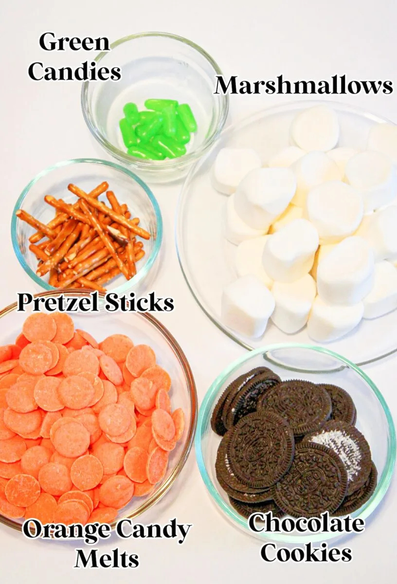 Five bowls, including a pumpkin patch marshmallow mix, green candies, pretzel sticks, orange candy melts, and chocolate cookies, are arranged on a white surface—each clearly labeled with its contents.