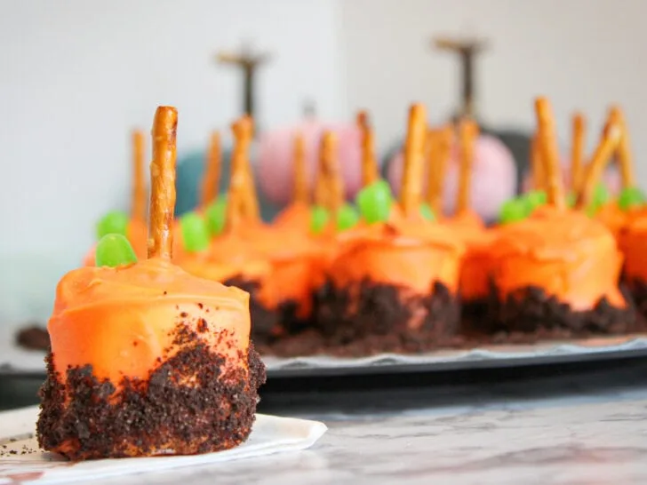 Marshmallows coated in orange candy with pretzel sticks and green candy on top, resembling pumpkins, are arranged on a tray to create a festive pumpkin patch marshmallow display.