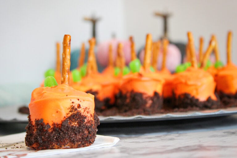 Marshmallows coated in orange candy with pretzel sticks and green candy on top, resembling pumpkins, are arranged on a tray to create a festive pumpkin patch marshmallow display.