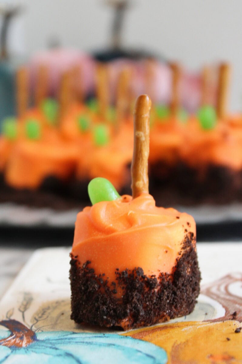 A small dessert with orange frosting, a chocolate cake base, a green candy, and a pretzel stick on top sits on a napkin—resembling a pumpkin patch marshmallow treat—with more desserts blurred in the background.