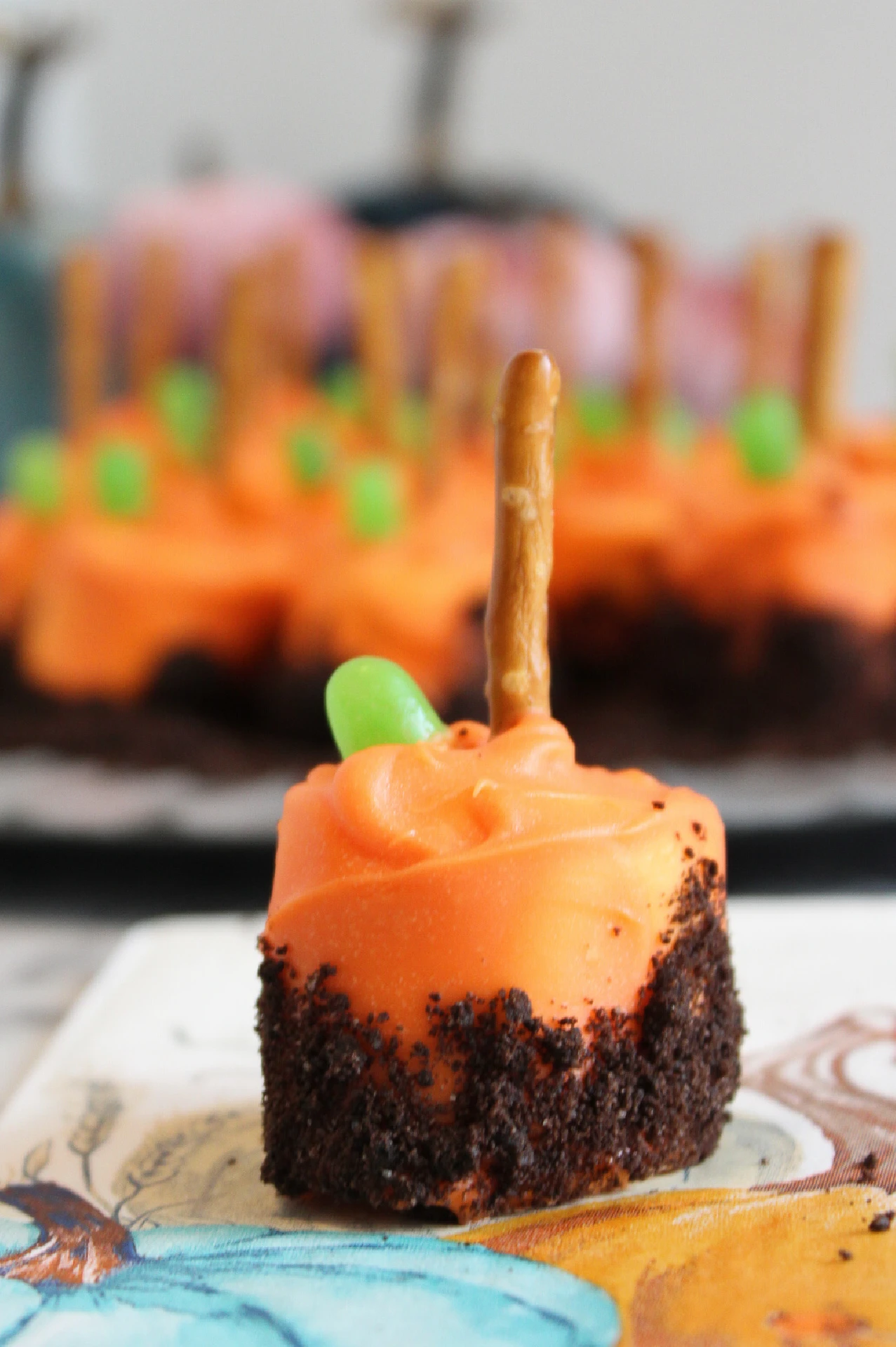 A small dessert with orange frosting, a chocolate cake base, a green candy, and a pretzel stick on top sits on a napkin—resembling a pumpkin patch marshmallow treat—with more desserts blurred in the background.
