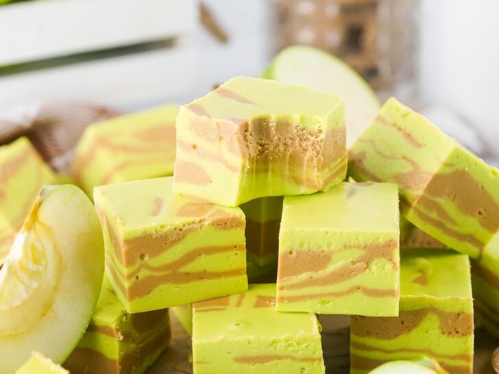 A stack of green and brown swirled caramel apple fudge pieces is displayed, with apple slices and a wooden crate in the background.