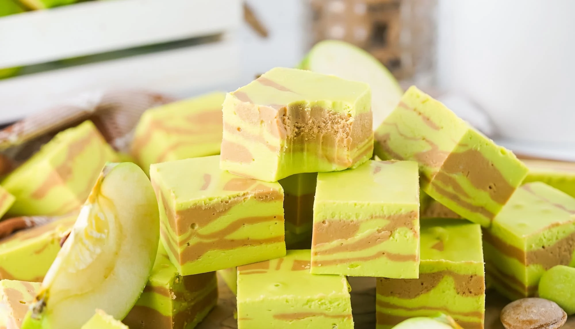 A stack of green and brown swirled caramel apple fudge pieces is displayed, with apple slices and a wooden crate in the background.