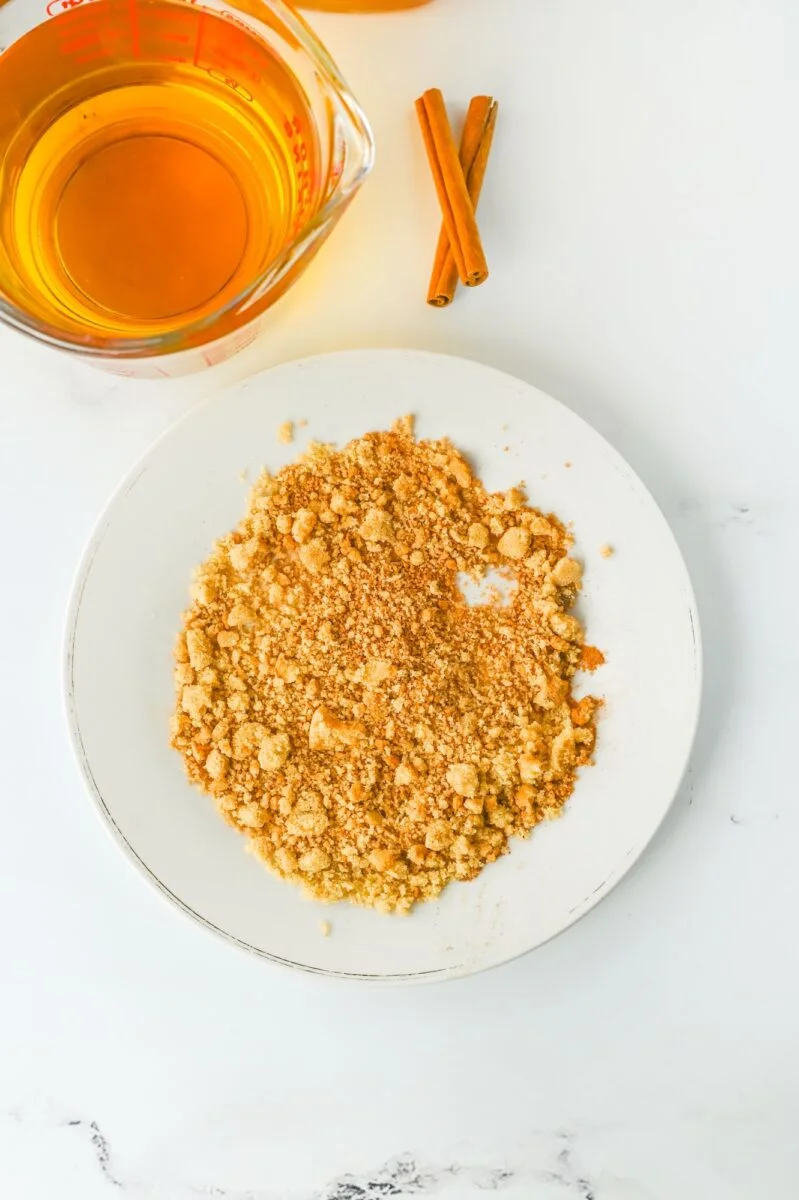 A white plate with a pile of crushed graham crackers, next to a measuring cup of amber apple pie cocktail and two cinnamon sticks on a white surface.