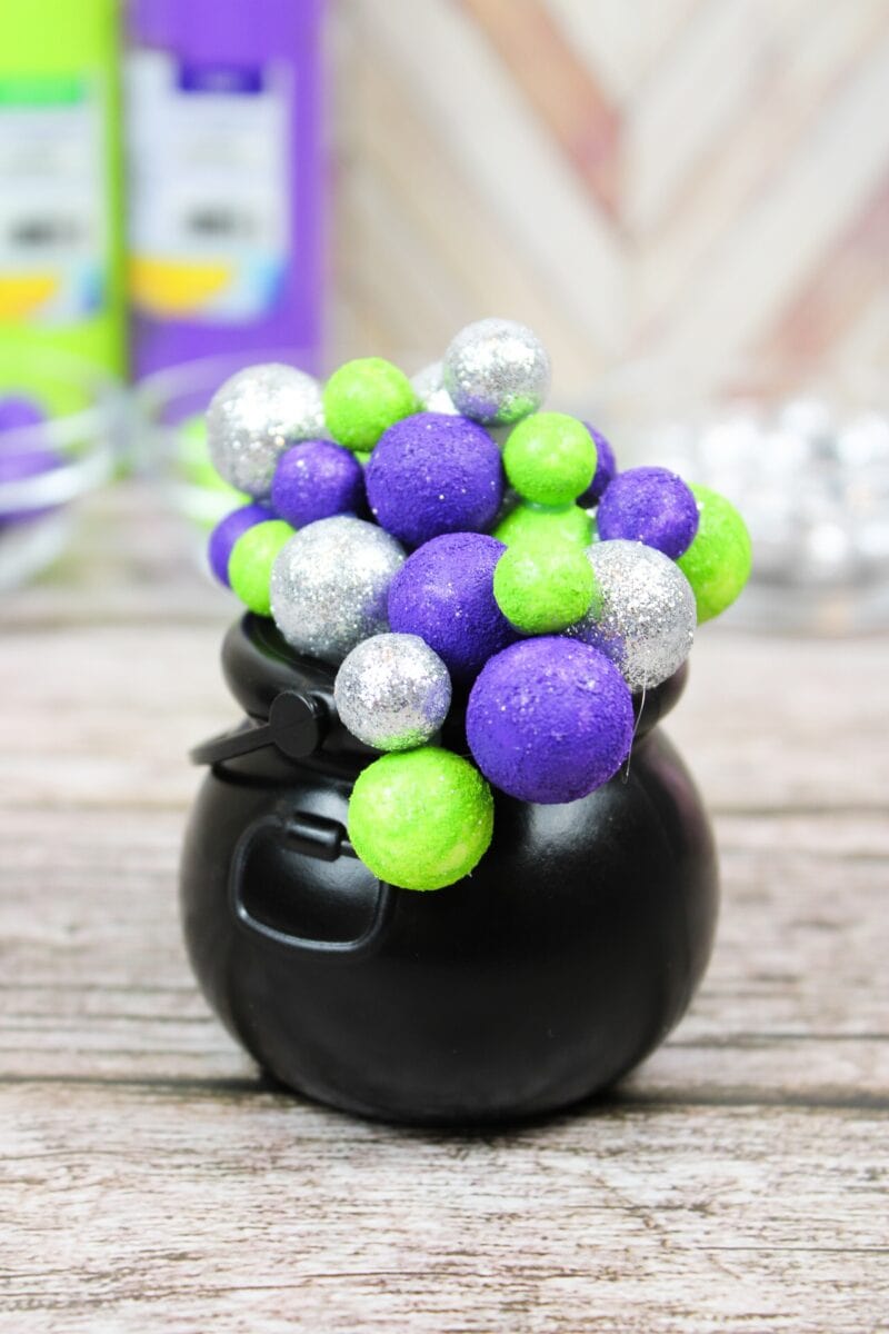 A small black cauldron filled with green, purple, and silver glittery foam balls, placed on a wooden surface.
