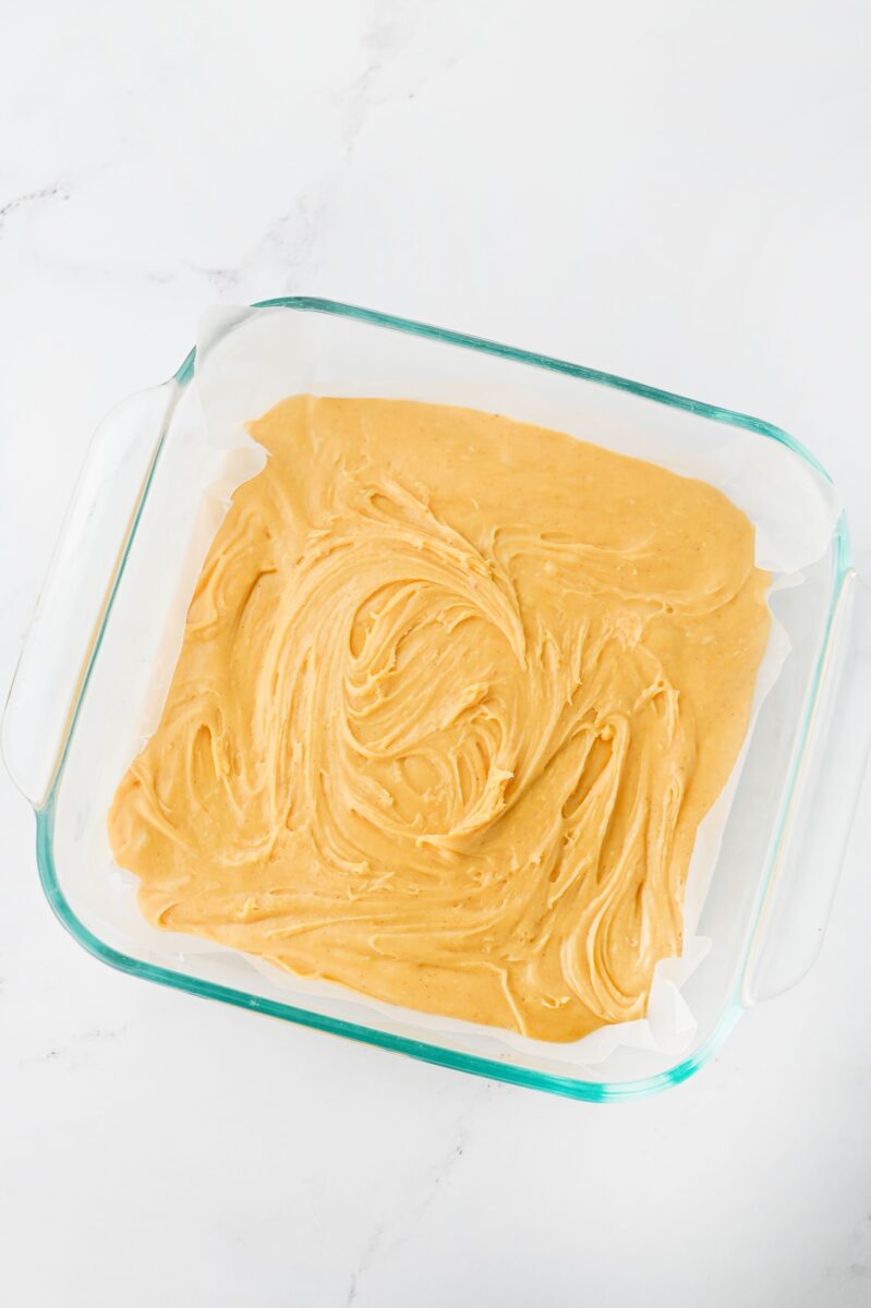 A glass baking dish lined with parchment paper and filled with swirled, light brown batter, placed on a white marble surface.