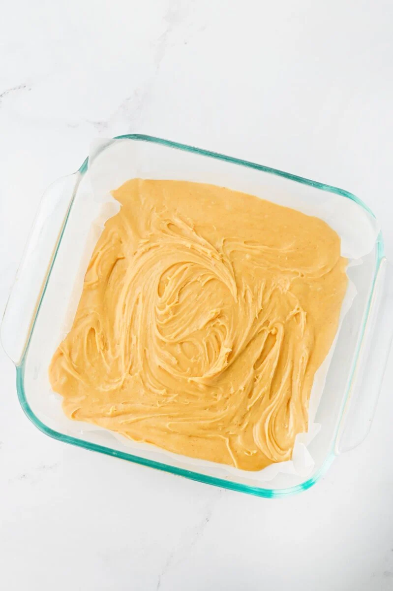 A glass baking dish lined with parchment paper and filled with swirled, light brown batter, placed on a white marble surface.