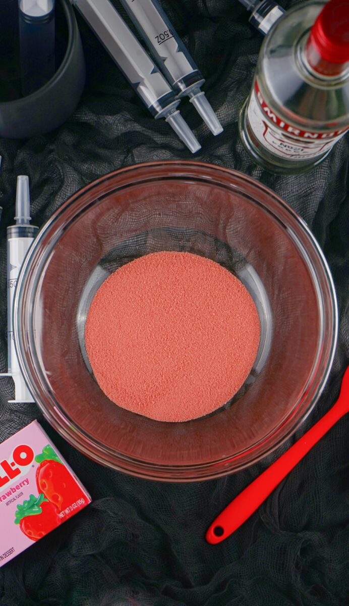 A glass bowl filled with strawberry Jell-O powder is surrounded by Jell-O packaging, syringe jello shots, a bottle of Smirnoff vodka, and a red spatula on a black surface.