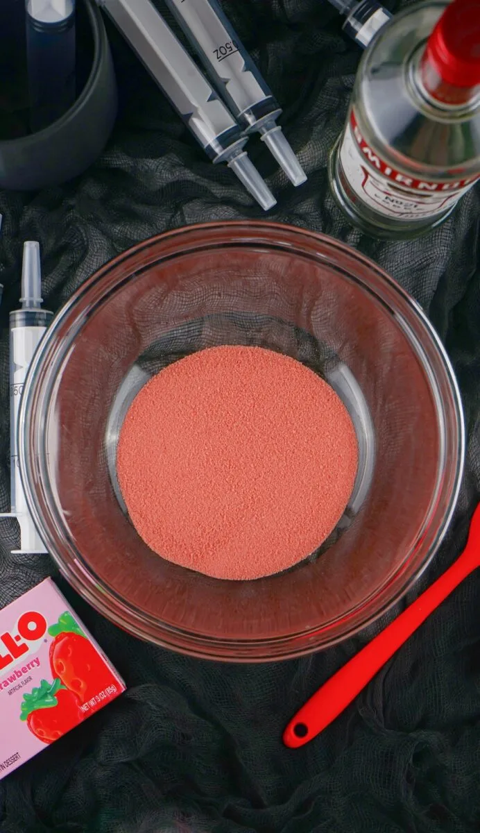 A glass bowl filled with strawberry Jell-O powder is surrounded by Jell-O packaging, syringe jello shots, a bottle of Smirnoff vodka, and a red spatula on a black surface.