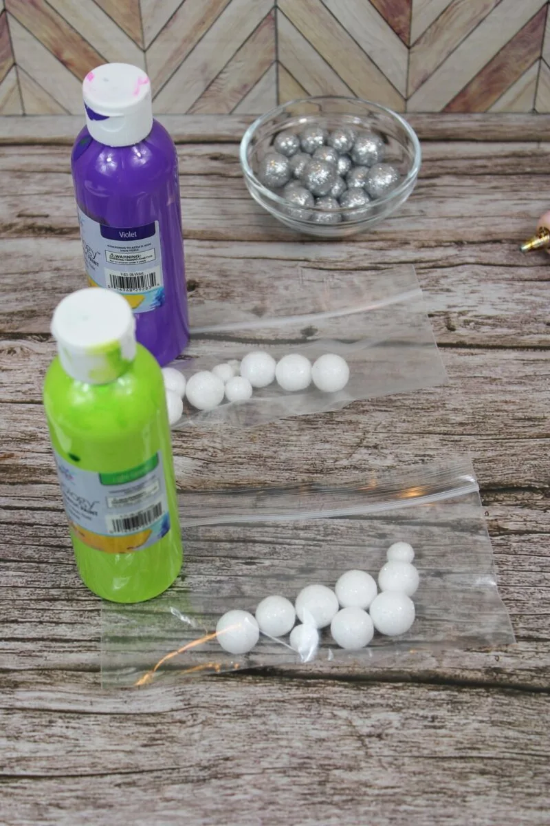 Two bottles of acrylic paint, a small bowl of silver beads, and two plastic bags containing white foam balls are arranged on a wooden surface.
