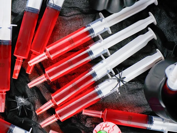 Several syringe jello shots filled with red liquid are arranged on a black net fabric, surrounded by Halloween-themed decorations including a fake spider and a plastic eyeball.