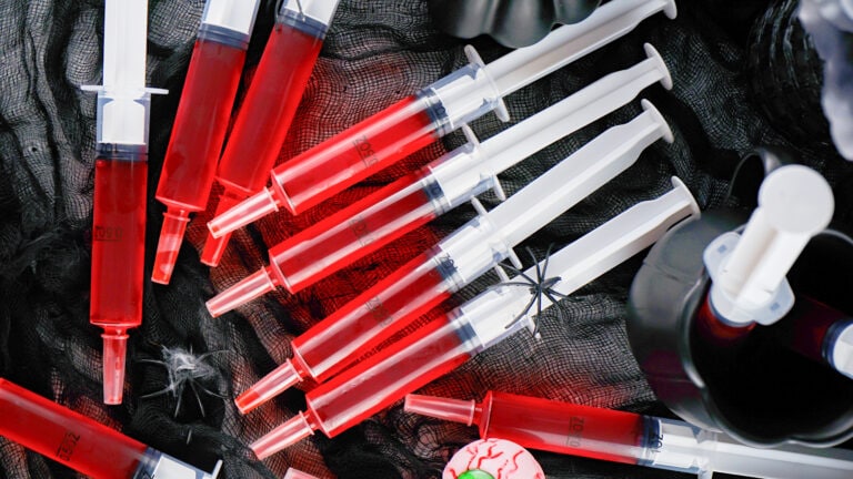 Several syringe jello shots filled with red liquid are arranged on a black net fabric, surrounded by Halloween-themed decorations including a fake spider and a plastic eyeball.