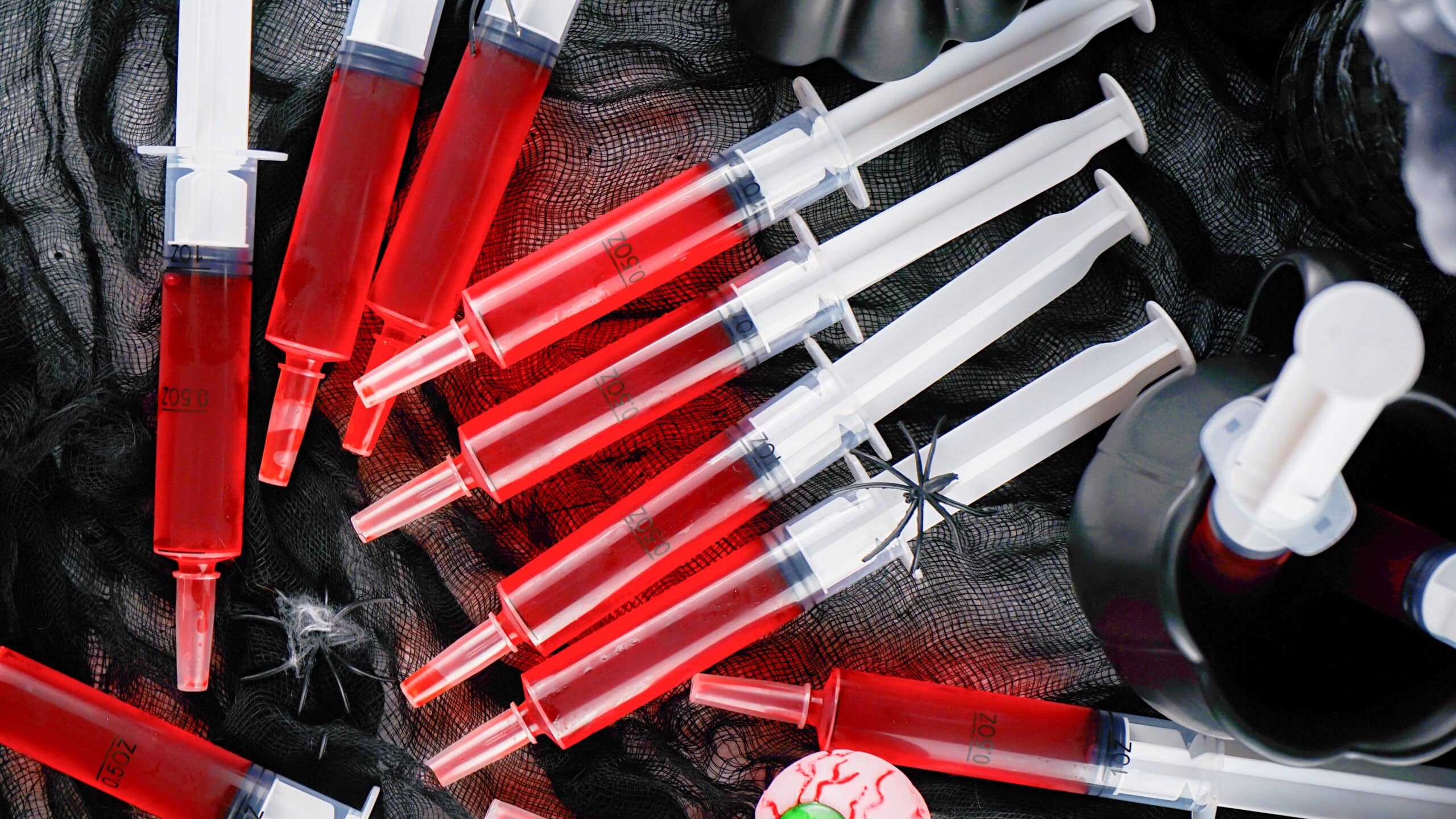 Several syringe jello shots filled with red liquid are arranged on a black net fabric, surrounded by Halloween-themed decorations including a fake spider and a plastic eyeball.
