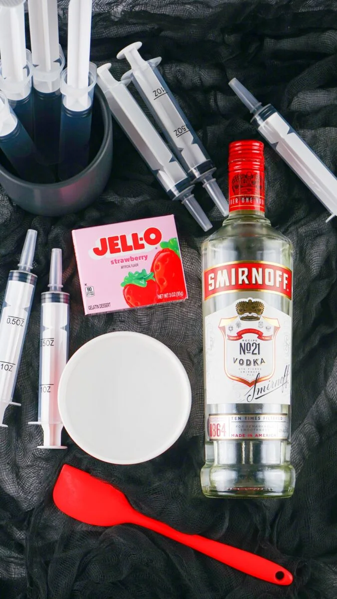 A bottle of Smirnoff vodka, a box of strawberry Jell-O, plastic syringes for syringe jello shots, a mixing bowl, and a red spatula are arranged on a black surface.