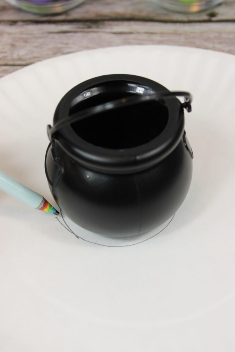 A small black plastic cauldron is placed upside down on a white paper plate, with a pencil tracing around its base.