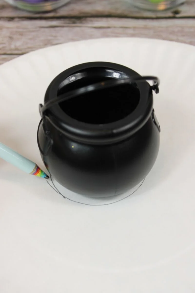 A small black plastic cauldron is placed upside down on a white paper plate, with a pencil tracing around its base.