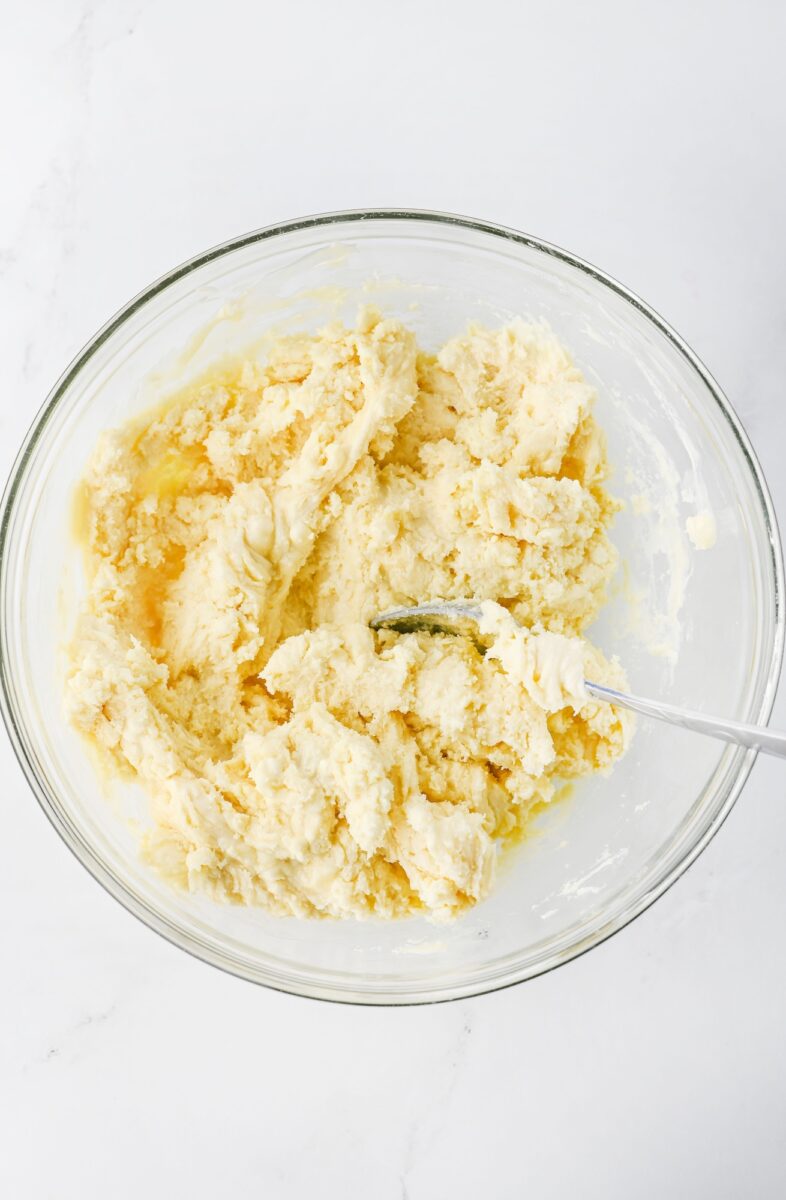 A glass bowl containing partially mixed dough with a metal spoon, set on a white surface.