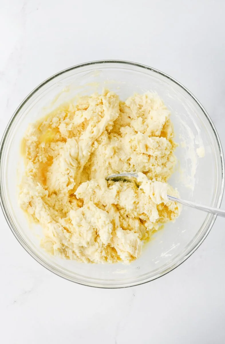 A glass bowl containing partially mixed dough with a metal spoon, set on a white surface.