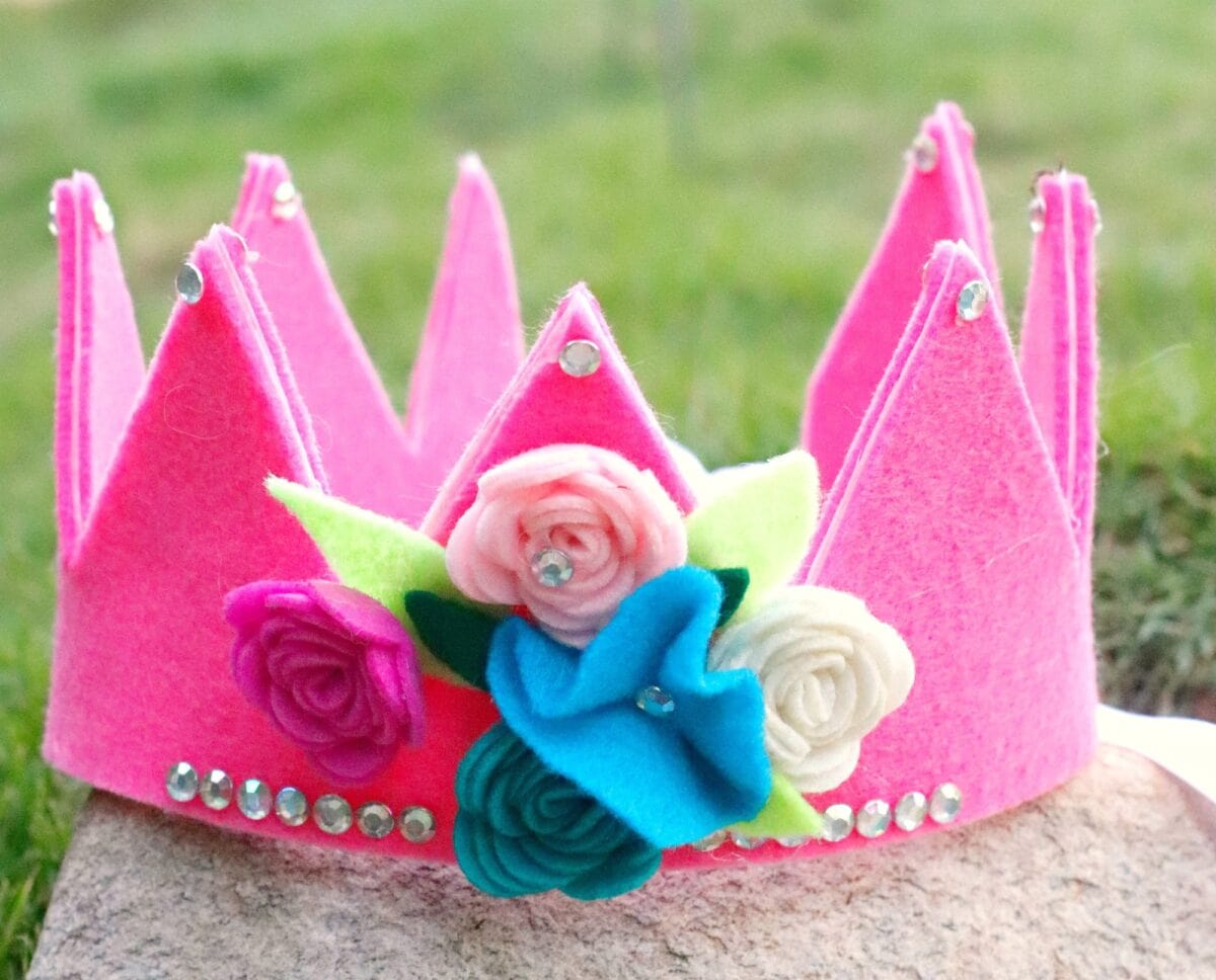 A pink felt crown decorated with faux flowers and rhinestones sits on a stone surface with grass in the background.
