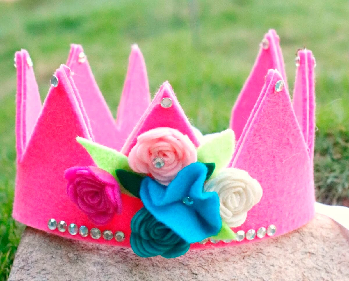 A pink felt crown decorated with faux flowers and rhinestones sits on a stone surface with grass in the background.