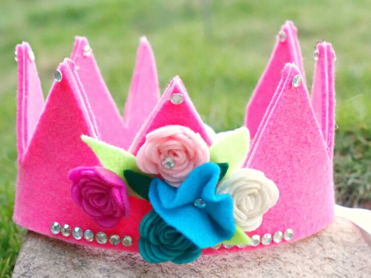 A pink felt crown decorated with felt flowers and rhinestones sits on a stone surface with grass in the background.