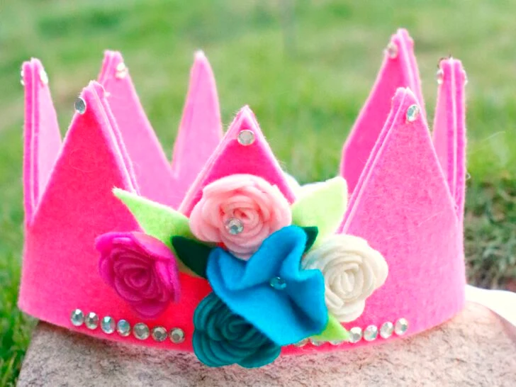 A pink felt crown decorated with felt flowers and rhinestones sits on a stone surface with grass in the background.