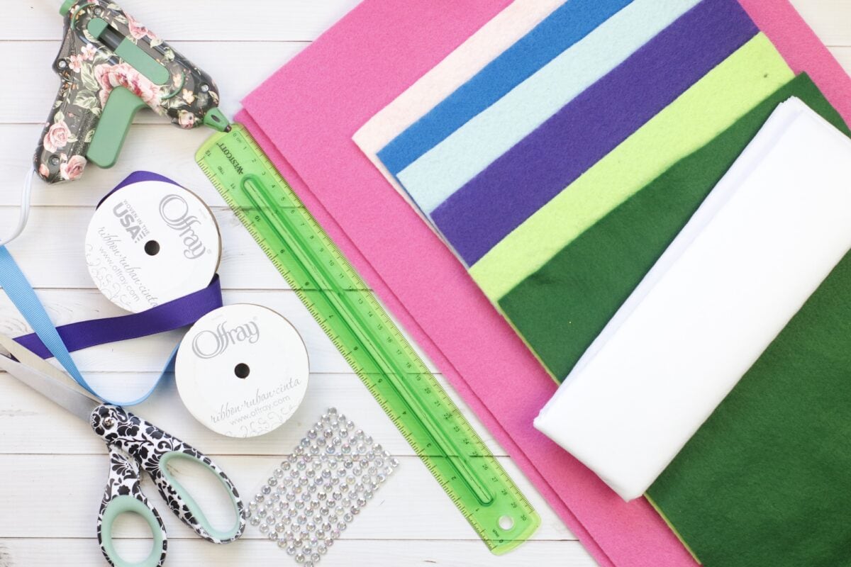 Assorted craft supplies including felt sheets, a green ruler, ribbons, a hot glue gun, scissors, and adhesive gems are arranged on a white surface.