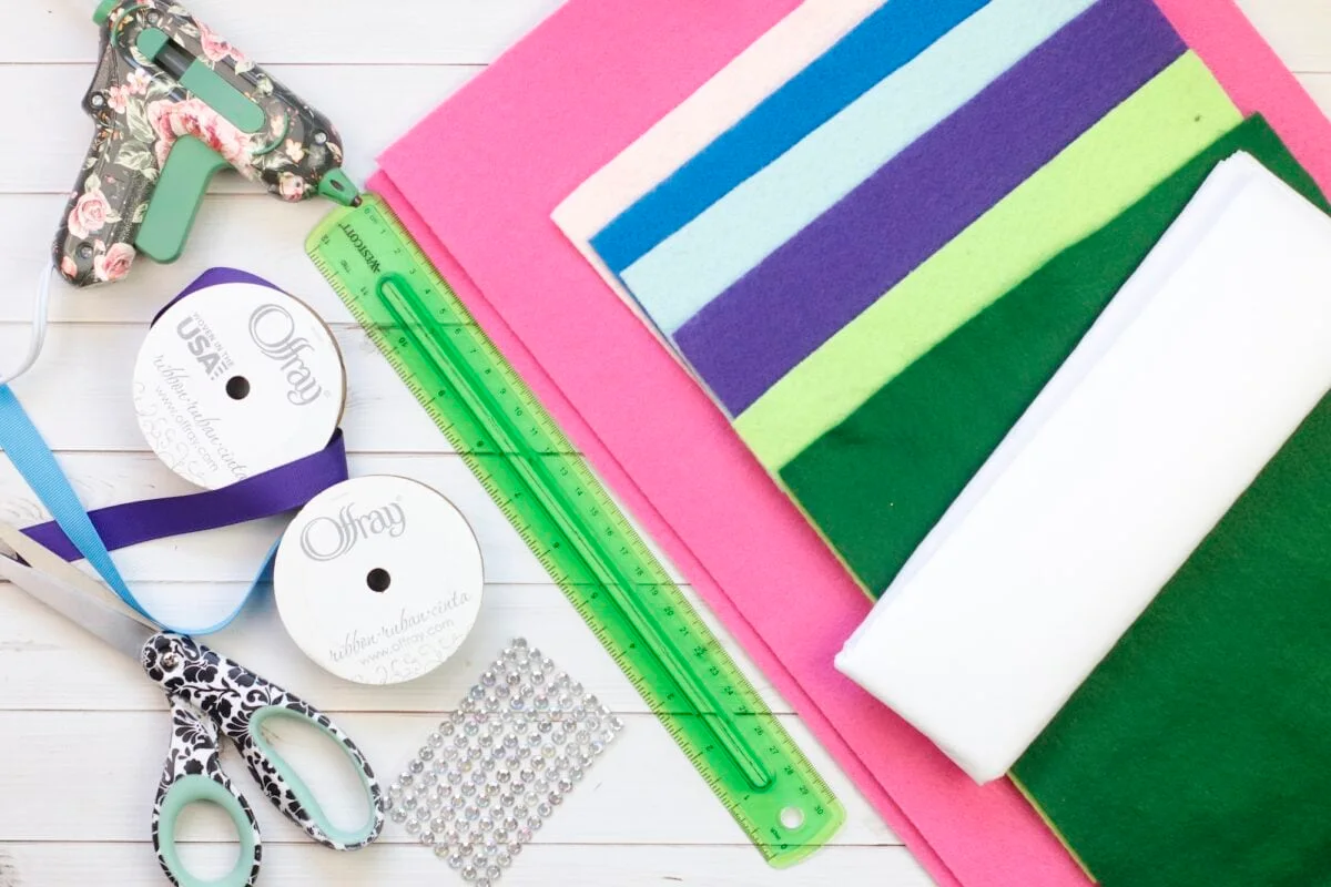 Assorted craft supplies including felt sheets, a green ruler, ribbons, a hot glue gun, scissors, and adhesive gems are arranged on a white surface.